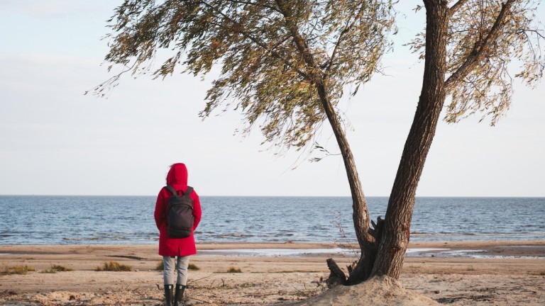 Woman in a red jacket stands under a tree and looks at the waves